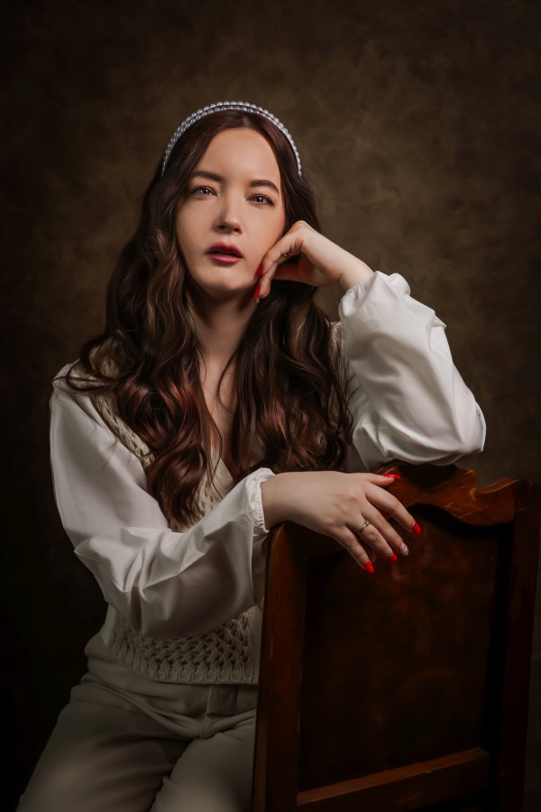 Fine art fotózás egy festményszerű stílus, a hölgy a barna széken ül, könyökölve, fehér és bézs ruhákban.