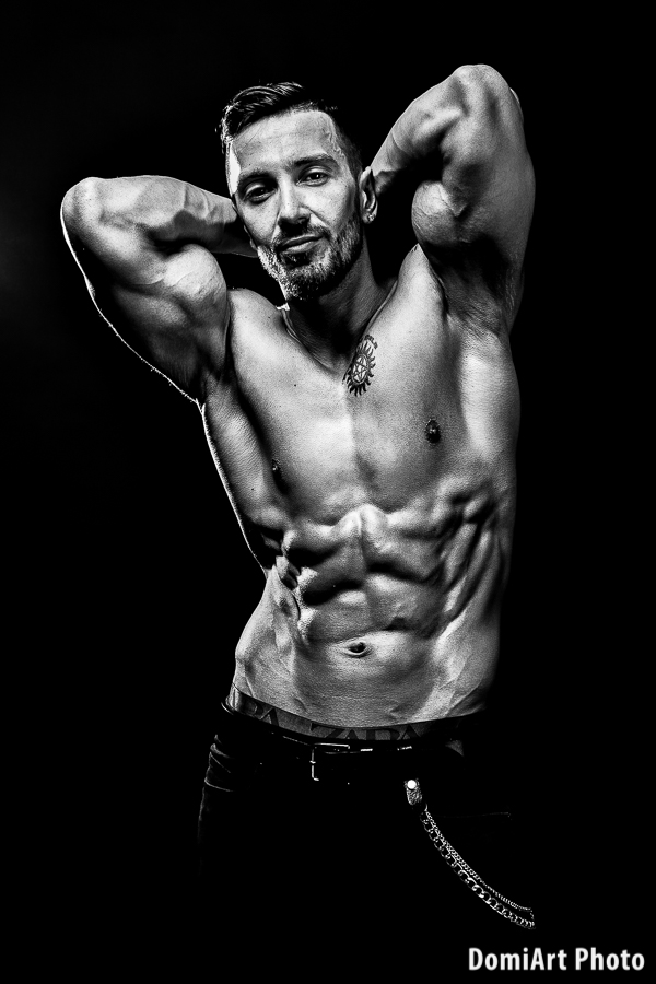 fekete-fehér képen férfi modell pózol, karjait a tarkóján tartja, így az izmok jobban látszanak. Férfi fitness fotózás
