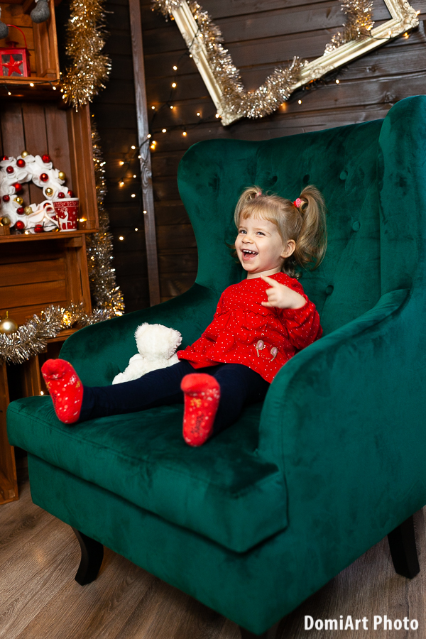 aranyos kislány piros ruhában, kétoldalra csurizott hajjal ül a hatalmas zöld fotelben - karácsonyi fotózás Debrecen