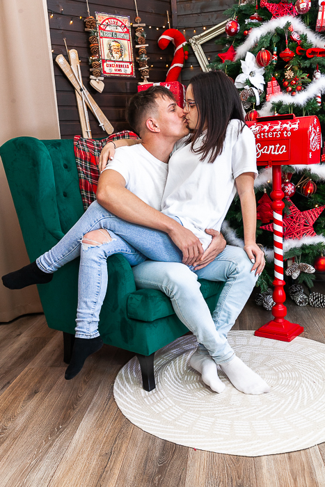 páros karácsonyi fotózás, a fiú ül a zöld fotelben, az ölében ül a lány és csókot adnak egymásnak