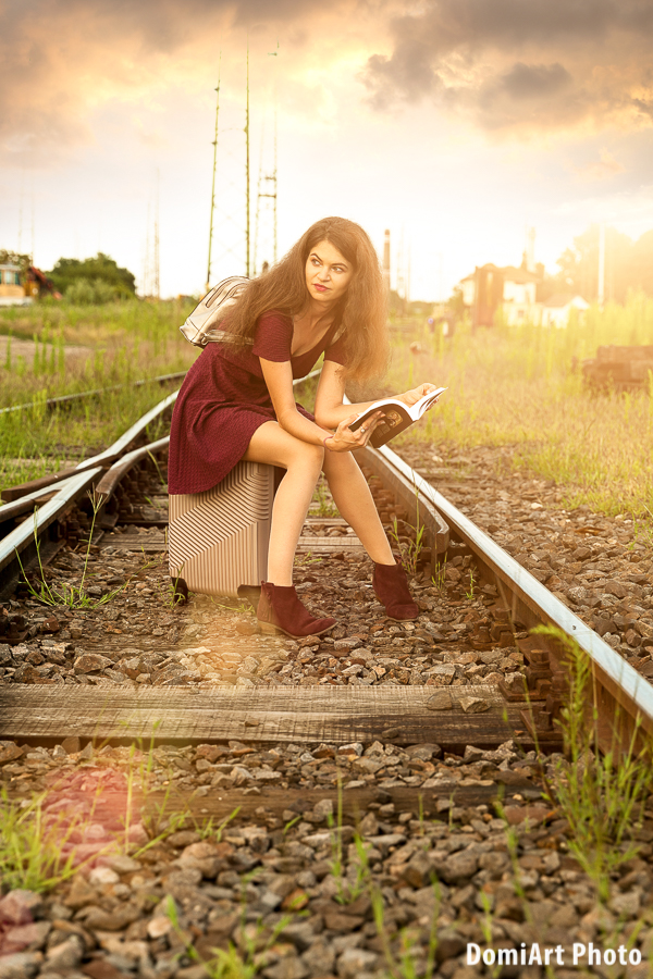 Vasútállomáson a sinen egy bőröndön ül egy lány, kezében könyvet tart - szabadtéri fotózás vasút állomáson